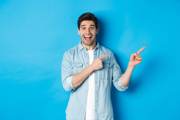 Obraz podekscytowanego przystojnego mężczyzny w swobodnym stroju, pokazującego reklamę, wskazującego palcami prosto na miejsce i uśmiechniętego, stojącego na niebieskim tle