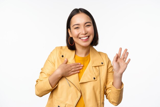 Obraz przyjaznej azjatyckiej dziewczyny w stylowym żółtym płaszczu, podnoszącej rękę, przedstawia się witającą machającą ręką, mówiącą „cześć”, stojącą na białym tle