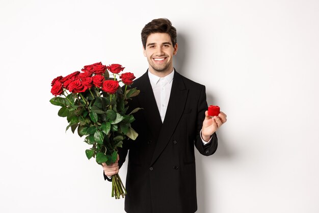 Obraz przystojnego mężczyzny w czarnym garniturze, trzymającego bukiet czerwonych róż i pierścionka, składającego propozycję, uśmiechniętego pewnie, stojącego na białym tle.