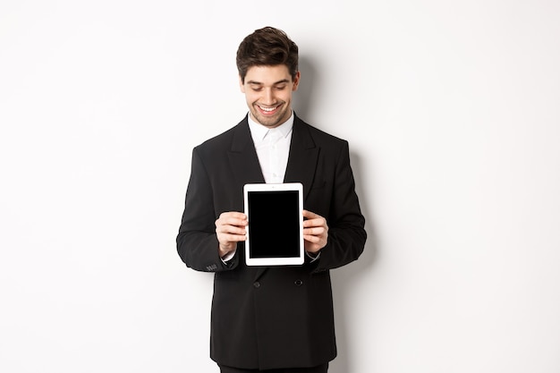 Obraz Przystojnego Przedsiębiorcy Płci Męskiej W Czarnym Garniturze, Patrzącego W Dół Na Ekran Cyfrowego Tabletu I Pokazującego Reklamę, Stojącego Na Białym Tle