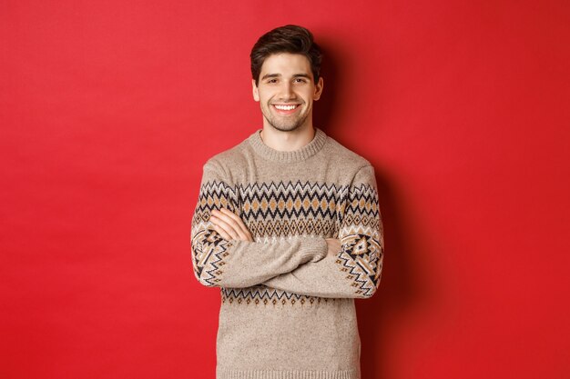 Obraz przystojnego szczęśliwego faceta w świątecznym swetrze, uśmiechającego się i patrzącego w kamerę, świętującego święta Bożego Narodzenia, stojącego na czerwonym tle
