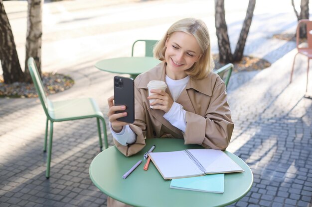 Obraz stylowej młodej studentki robiącej selfie w kawiarni na ulicy, pozując z ulubionym napojem