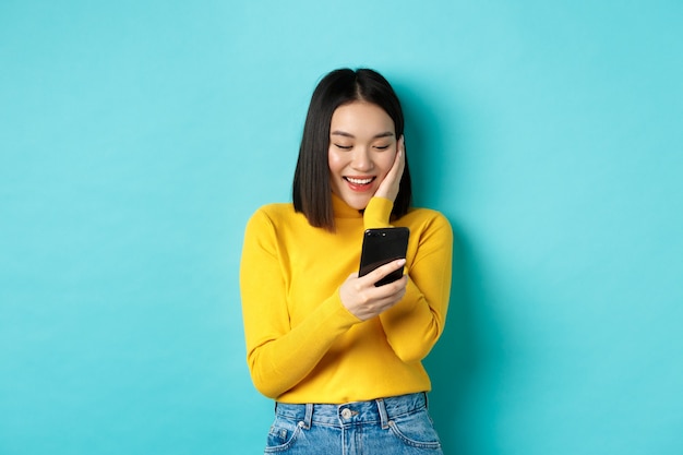 Obraz szczęśliwej azjatyckiej kobiety czytającej wiadomość na ekranie telefonu komórkowego i uśmiechającej się, rozmawiającej w aplikacji na smartfona, stojącej na niebieskim tle
