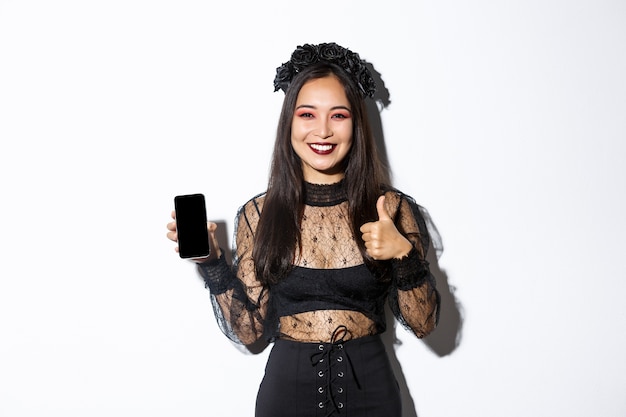 Obraz szczęśliwej i zadowolonej Azjatki w kostiumie na halloween pokazujący kciuki do góry i pokazujący ekran telefonu komórkowego, uśmiechnięty zadowolony, stojący na białym tle.