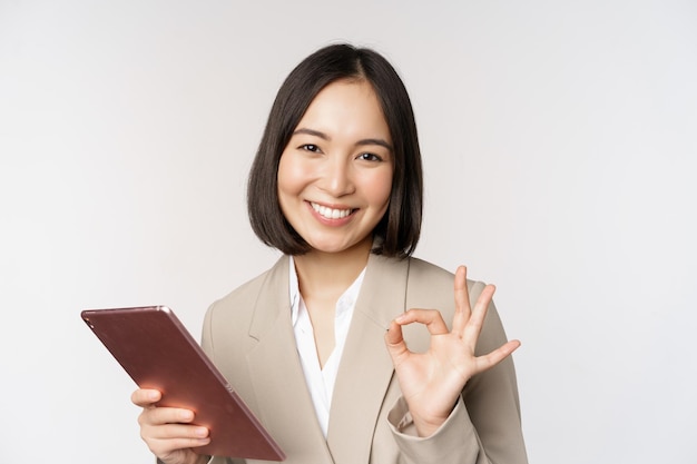 Obraz udanej azjatyckiej bizneswoman trzymającej cyfrowy tablet pokazujący znak ok, zapewniający klienta stojącego na białym tle