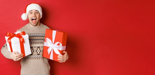 Bezpłatne zdjęcie obraz wesołego, atrakcyjnego faceta trzymającego prezenty świąteczne, stojącego w santa hat i zimowego swetra, uśmiechającego się zdziwionego, stojącego na czerwonym tle.