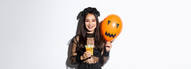 Obraz wesołej azjatyckiej dziewczyny sztuczki lub leczenia na Halloween trzymającej słodycze i pomarańczowy balon z s