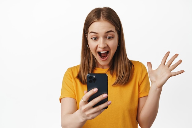 Obraz zaskoczonej szczęśliwej kobiety wzdycha, czytając ekran smartfona i krzycząc podekscytowany, krzycząc z radości, odbieraj pozytywne wiadomości na telefonie komórkowym, stojąc na białym tle