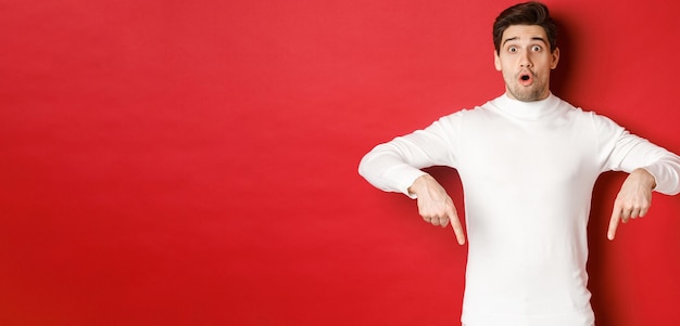 Obraz zdziwionego, atrakcyjnego faceta w białym swetrze, pokazującego ogłoszenie, wskazującego palcami w dół i wyglądającego na zdziwionego, stojącego na czerwonym tle