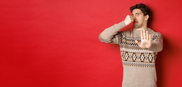 Obraz zniesmaczonego przystojnego mężczyzny w zimowym swetrze, odrzucającego coś z nieprzyjemnym zapachem, zamkniętego nosa i podnoszącego rękę, by odmówić stojącego na czerwonym tle