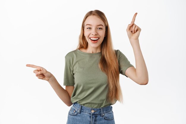Obraz zrelaksowanej, szczęśliwej młodej kobiety wskazującej palcami na boki u góry i w lewo, uśmiechniętej i śmiejącej się, polecającej dwa produkty, pokazującej możliwości wyboru, stojącej na białym tle