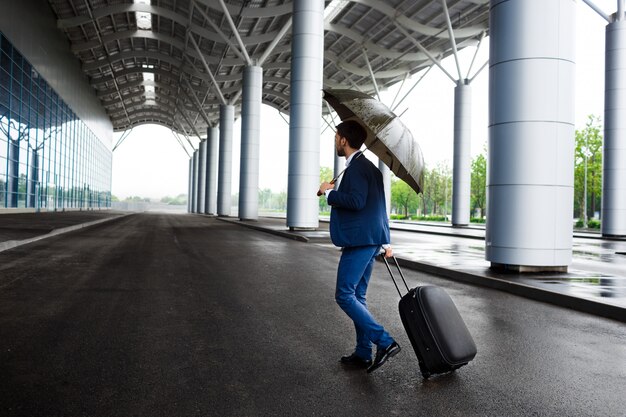 Obrazek młodego biznesmena mienia walizka i parasol przy dżdżystym terminal