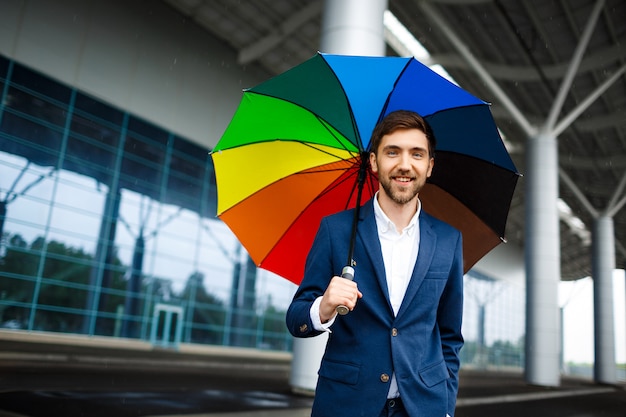 Obrazek młody rozochocony biznesmen trzyma pstrobarwnego parasol w dżdżystej ulicie