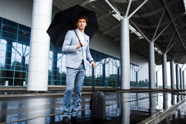 Obrazek ufny młody redhaired biznesmen trzyma czarną walizkę w deszczu przy terminal i parasol