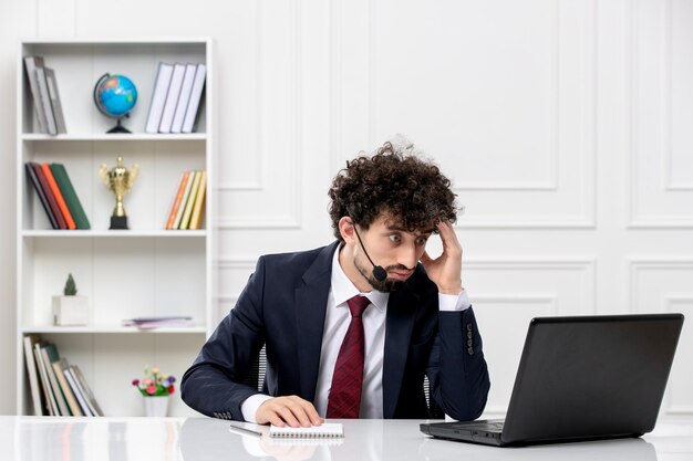 Obsługa klienta kędzierzawy brunetka młody mężczyzna w garniturze biurowym z laptopem i zestawem słuchawkowym dotykającym głowy