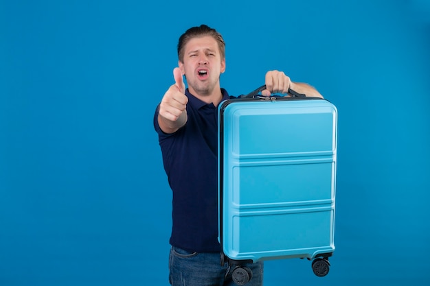 Oburzony młody przystojny podróżnik mężczyzna trzyma walizkę wskazując palcem na aparat niezadowolony i sfrustrowany stojąc nad błękitną przestrzenią