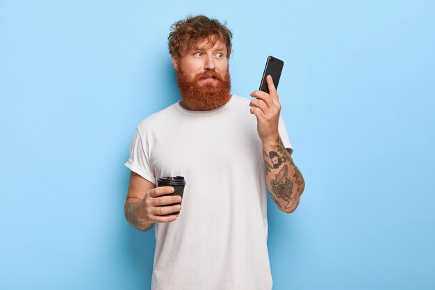 Oburzony, zdziwiony rudowłosy facet pozujący ze swoim telefonem