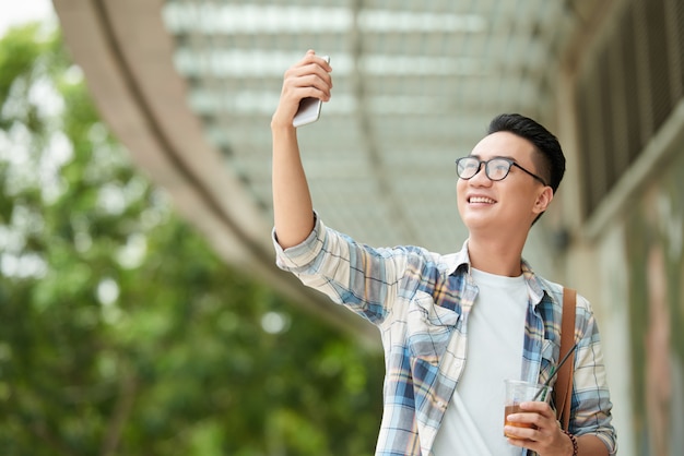 Od niechcenia ubrany mężczyzna Azji idąc przez centrum handlowe z napojem i biorąc selfie na smartfonie