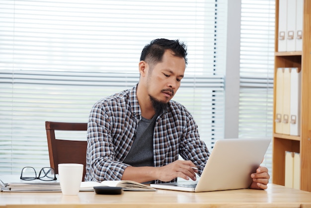 Od niechcenia ubrany mężczyzna filipiński siedzi w biurze i pracuje na laptopie