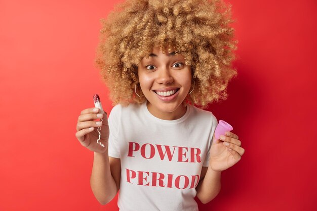 Od pasa w górę strzał pięknej wesołej kobiety trzyma używany tampon i kubek menstruacyjny do ochrony przed wyciekiem, ubrany w luźną koszulkę na białym tle nad żywym czerwonym tłem Produkty do higieny intymnej