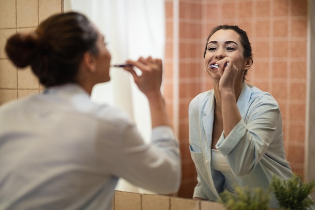 Odbicie w lustrze młodej uśmiechniętej kobiety szczotkującej zęby w łazience