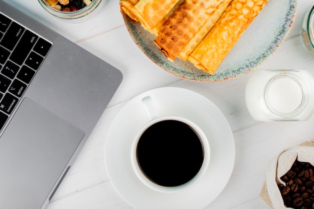 Odgórny widok filiżanka kawy z opłatkowymi rolkami i laptopem na białym drewnianym tle