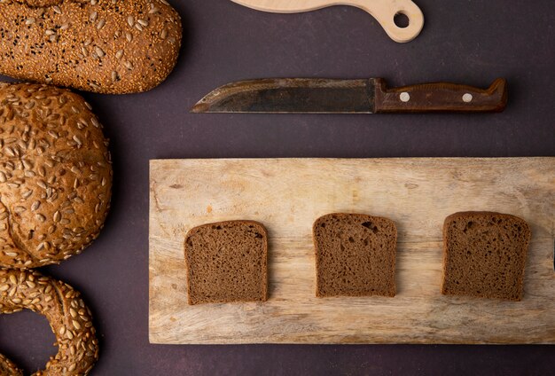 Odgórny widok żyto chleba plasterki na tnącej desce z chlebami i nożem wokoło na bordowym tle