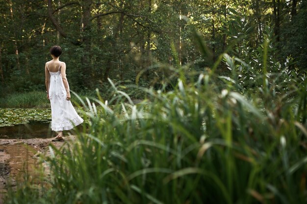 Odkryty letni obraz romantycznej uroczej młodej kobiety noszącej długą białą sukienkę, relaksującej się samotnie w dzikiej naturze w weekend, stojącej nad stawem w tle ze świeżą zieloną trawą na pierwszym planie