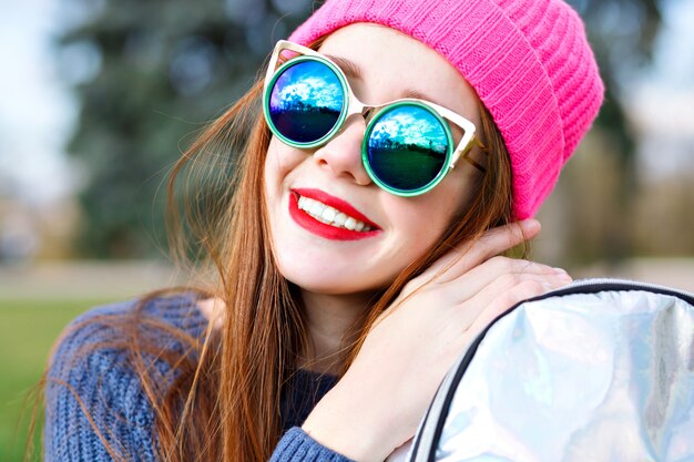 Odkryty pozytywny portret mody oszałamiającej pięknej rudej kobiety, pozuje w parku miejskim, w kapeluszu hipster i pogodzie, modne luksusowe okulary przeciwsłoneczne, młodzież, wakacje, czerwone usta, pastelowe kolory.