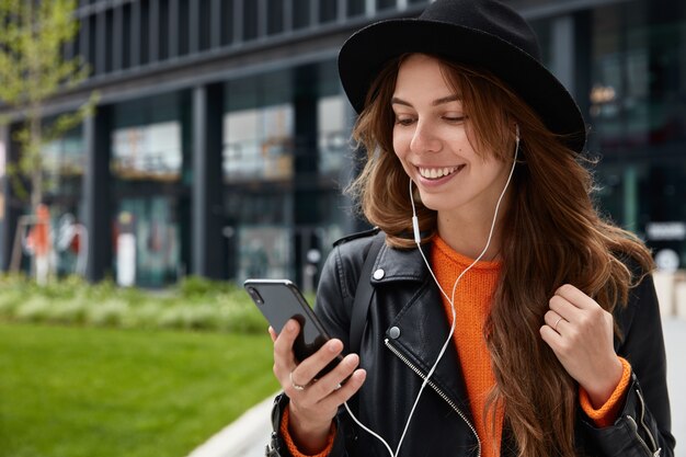 Odkryty strzał kaukaskiej kobiety lubi słuchać ścieżki dźwiękowej, używa nowoczesnego telefonu komórkowego i słuchawek, pozuje w centrum miasta, ma zębaty uśmiech