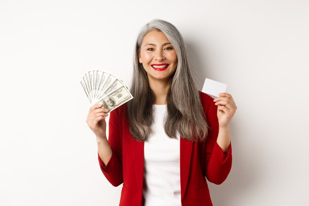 Odnosząca sukcesy azjatycka starsza bizneswoman pokazująca pieniądze w dolarach i plastikowej karcie, uśmiechnięta szczęśliwa do kamery, ubrana w czerwoną marynarkę i makijaż