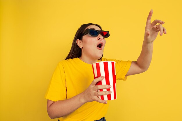 Oglądanie kina w okularach 3D z popcornem. Kaukaski kobieta na żółtym tle studio. Piękny model brunetka w stylu casual. Pojęcie ludzkich emocji, wyraz twarzy, sprzedaż, reklama, copyspace.