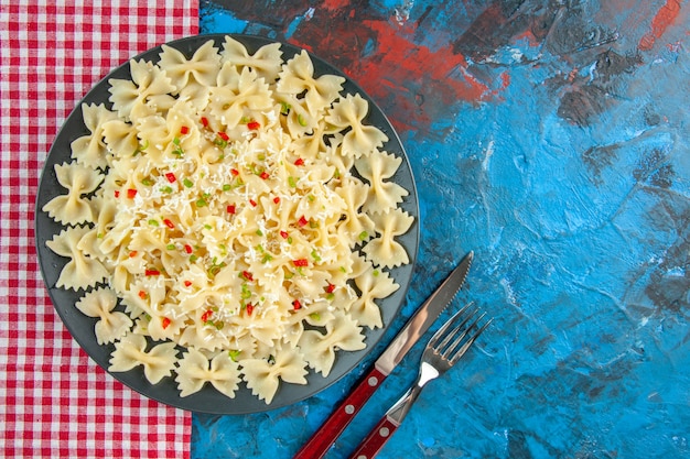 Ogólny widok surowego włoskiego makaronu farfalle z warzywami na czerwonym ręczniku w paski obok sztućców ustawionych na niebieskim stole