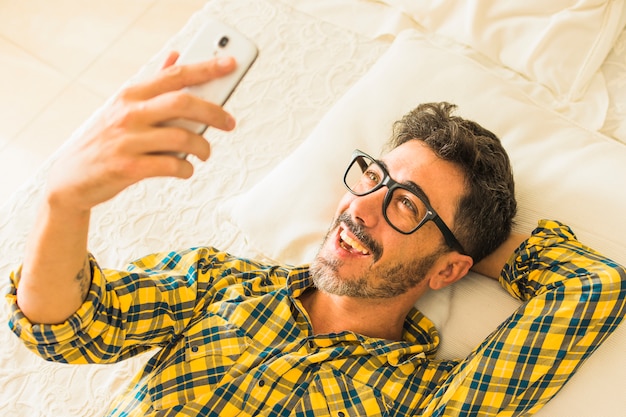 Ogólny widok uśmiechniętego człowieka leżącego na łóżku patrząc na smartphone