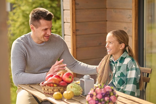 Ojcostwo. Uśmiechnięty młody dorosły mężczyzna w zwykłych ubraniach rozmawiający dotykając ramienia z zainteresowaniem słuchający długowłosej córki siedzącej przy stole na otwartej werandzie w pogodne popołudnie