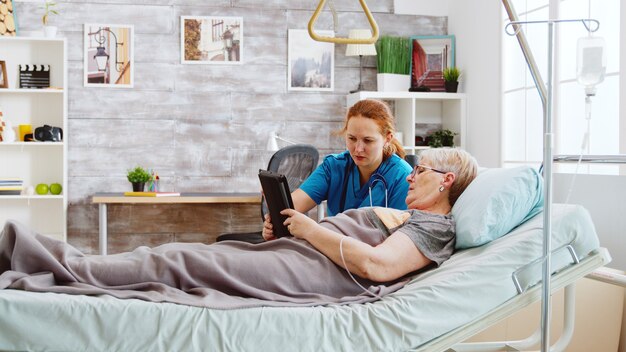 Opiekunka pomaga starej niepełnosprawnej kobiecie leżącej w szpitalnym łóżku w korzystaniu z cyfrowego tabletu. Jasny pokój z dużymi oknami