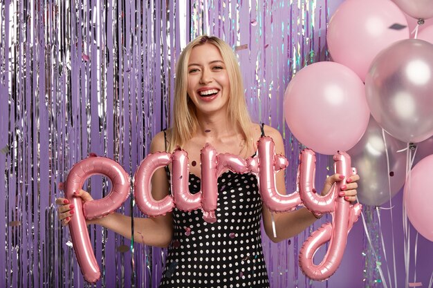 Optymistyczna szczęśliwa blondynka nosi modną sukienkę w groszki, robi zdjęcie z balonami na imprezie