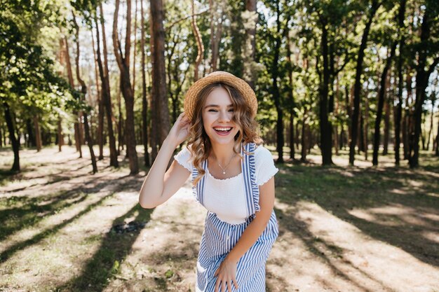 Oszałamiająca dziewczyna w modnym stroju uśmiechnięta podczas sesji zdjęciowej w lesie. Urocza modelka w kapeluszu, ciesząc się dobrym dniem w parku.