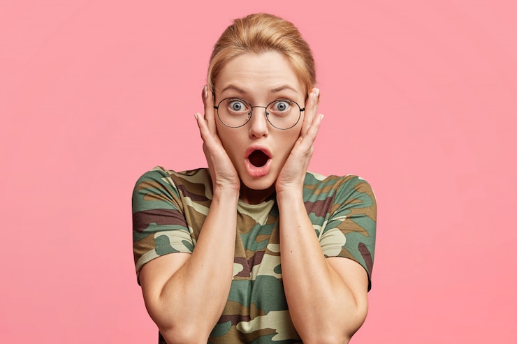 Bezpłatny plik Zdjęcie oszołomiona blond modelka w okularach, zdumiona czymś, trzyma usta otwarte, ubrana w koszulkę kamuflażową, odizolowaną na różowo
