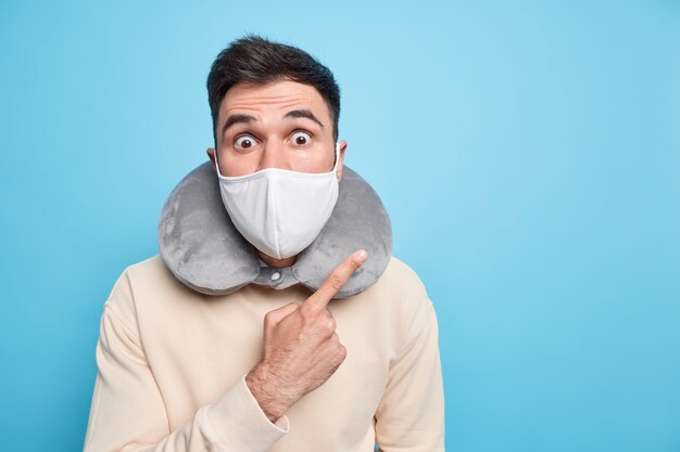 Oszołomiony emocjonalnie dorosły mężczyzna nosi maskę ochronną podczas epidemii koronawirusa podaje zalecenia, jak zachować bezpieczeństwo, prosi o przestrzeganie zasad kwarantanny, nosi poduszkę pod szyję z dala od miejsca kopiowania