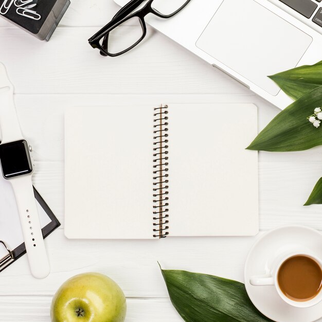 Otwarty notatnik spiralny otoczony papierami biurowymi, jabłkiem i eleganckim zegarkiem na biurku