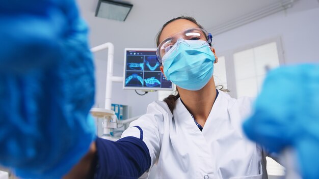 Pacjent z punktu widzenia dentysty w masce ochronnej trzymającej narzędzia badający osobę z bólem zęba siedzącą na fotelu stomatologicznym, podczas gdy pielęgniarka przygotowuje narzędzia do zabiegu.
