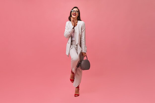 Pani w garniturze, śmiejąc się i trzymając torbę na różowym tle. Piękna biznesowa kobieta w okularach iz czerwoną szminką pozowanie na kamery.