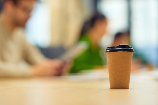 Papierowy kubek do kawy stojący na stole w biurze z osobami pracującymi w tle