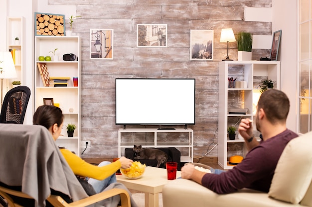 Para patrząca na odizolowany ekran telewizora w przytulnym salonie podczas jedzenia jedzenia na wynos