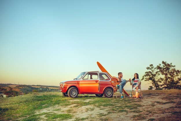 Para siedzi i odpoczywa na plaży, grając na gitarze w letni dzień, w pobliżu rzeki. Miłość, szczęśliwa rodzina, wakacje, podróże, koncepcja lata.