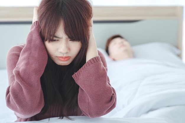 Para w łóżku mężczyzna śpi i chrapie, podczas gdy kobieta nie może spać, zamykając uszy dużymi poduszkami
