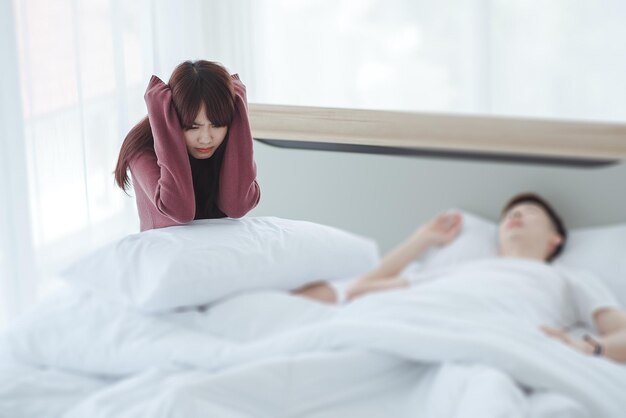 Para w łóżku mężczyzna śpi i chrapie, podczas gdy kobieta nie może spać, zamykając uszy dużymi poduszkami