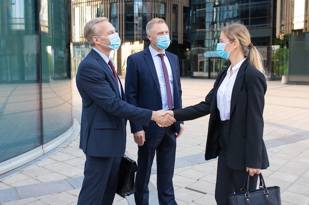 Partnerzy biznesowi w maskach na twarz robią interesy lub witają się. Profesjonalny sukces bizneswoman i biznesmeni stojący na zewnątrz i uścisk dłoni. Koncepcja negocjacji, ochrony i partnerstwa