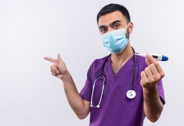 Patrząc do kamery młody lekarz płci męskiej ubrany w fioletową odzież chirurga i maskę medyczną stetoskop trzyma punkty termometru z boku na na białym tle